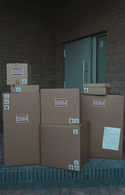 Several large cardboard boxes and smaller parcels stacked outside a building near a blue patio door, with some boxes marked 'Fragile' and labeled with handling symbols, indicating they contain delicate items for home relocation. The boxes are placed on a brick-paved area adjacent to a brick wall, with some positioned on the ground and others slightly elevated. The scene suggests the packing stage of a moving process, with packaging materials visible and prepared for transportation. This setup is typical of house removal services provided by firms like Man and Van Eltham during the furniture transport and packing phases of a move, ensuring careful handling of valuables during a home relocation.