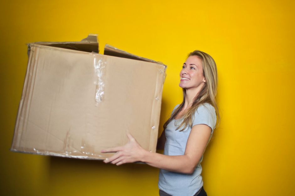A woman with long blonde hair, wearing a light grey T-shirt, is standing indoors against a bright yellow wall. She is smiling while holding a large, partially unfolded cardboard box, which is secured with clear packing tape. The box appears to be filled with or ready for packing household items, indicative of a moving or home relocation process. Behind her, part of a doorway and a glimpse of the floor are visible. The scene suggests the packing stage of a house move, with the woman preparing items for transport. This image aligns with professional removals and packing services offered by Man and Van Eltham, emphasizing efficient packing and furniture transport during house removals.