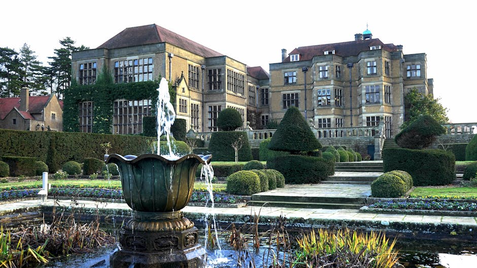 View of a historic stone mansion with multiple bay windows and a red tiled roof, situated behind a formal garden with trimmed bushes, tiered stone steps, and flowerbeds. In the foreground, a decorative fountain with water cascading from a large, ornate metal basin is positioned in a pond surrounded by dried aquatic plants. The garden features symmetrical, sculpted evergreen and deciduous shrubs, with a paved pathway leading towards the mansion. The scene suggests a quiet, well-maintained estate in daylight, with no visible furniture or moving equipment, reflecting a setting suitable for house removals or a home relocation involving careful planning and logistics by specialists such as Man and Van Eltham.