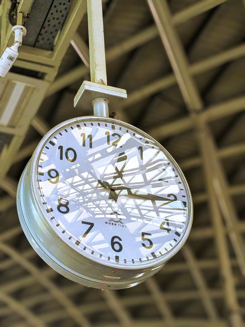 A large round wall clock with a white face, black numerals, and black hour and minute hands, hangs from a metal support beneath a high, industrial-style ceiling with exposed beams and metal framework. The clock displays the time as approximately 4:09. The ceiling appears to be inside a covered outdoor or railway station area, with natural lighting filtering through. This setting indicates a public space such as a station or loading area used in home relocation or furniture transport processes. Man and Van Eltham, specializing in removals, may utilize such spaces during the loading or unloading stages of house moves, involving careful handling of furniture or boxes seen nearby. The clock's position and visible surroundings highlight the importance of timing and logistics in efficient moving operations.
