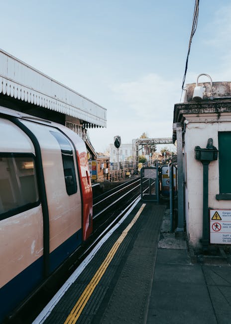 View of a train arriving at Eltham station platform with a man and van parked nearby, which is partially visible on the right side of the image. The platform features tactile paving strips for accessibility, with several cardboard boxes, wrapped furniture, and packing materials stacked near the edge, ready for loading onto the vehicle. The loading process appears to involve careful handling of household items as part of a home relocation or furniture transport. In the background, railway tracks extend into the distance, with station signage and infrastructure such as overhead electrical lines and a small building with a green window. The setting is outdoors during daytime with clear weather, and the scene captures a moment of organized loading and packing for a house move, with Man and Van Eltham providing professional removal services to support the relocation process.